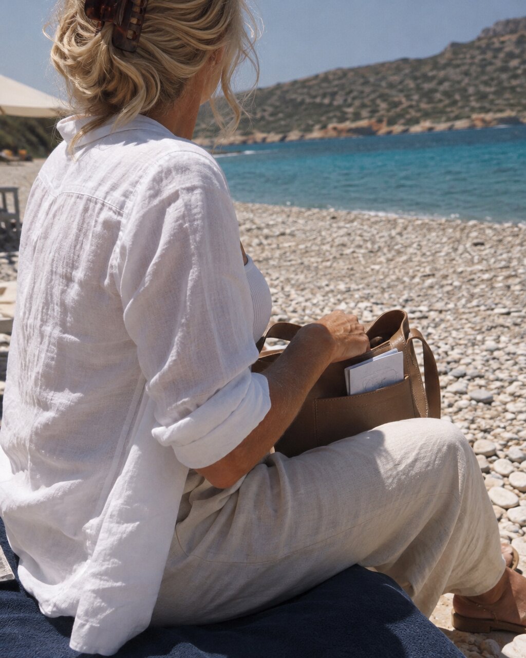 Woman seen from behind in white linen, sitting on a pebble beach facing the sea